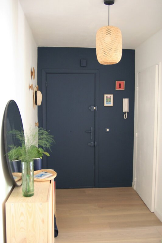 Porte et mur d'entrée bleu nuit. Suspension en osier. Cadres colorés. Console et miroir posé. Vase et vide poche.