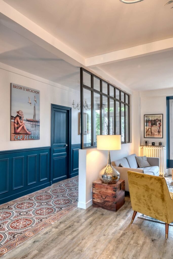 Vue salon et entrée séparée par une verrière atelier en bois. Porte et demi-mur peint en bleu. Canapé beige et fauteuil velours jaune. Carrelage ancien rouge et blanc au sol.