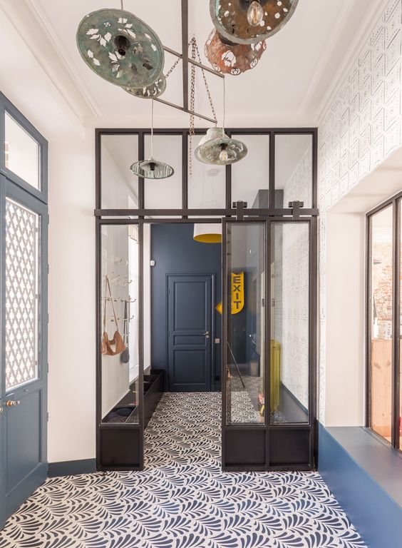 Entrée avec porte et mur bleu foncé. Verrière atelier noire avec porte vitrée coulissante. Carrelage au sol bleu et blanc motifs végétaux.
