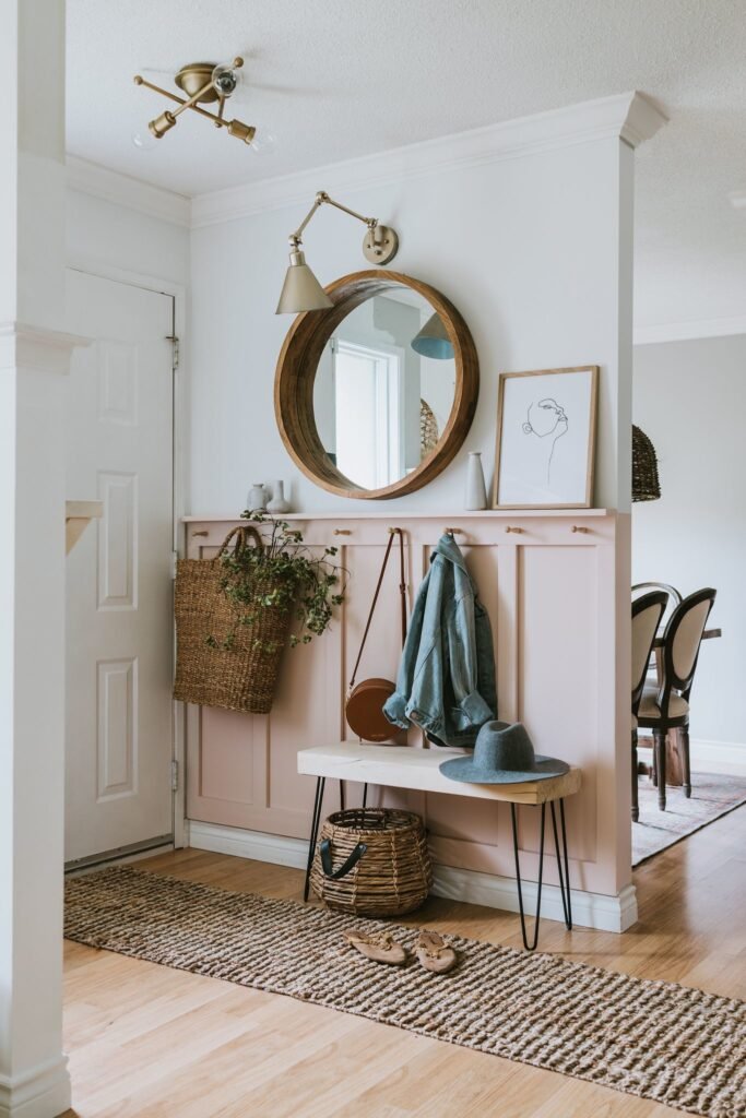 Bien décorer son entrée. Entrée avec boiserie rose. Patères. Miroir rond en bois. Banquette en bois. Panier avec fleurs. Tapis.